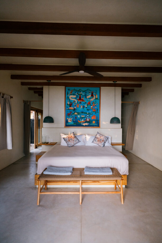 Spacious, serene bedroom at Samas Hotel La Saladita with a king-size bed, exposed wooden beams, and a vibrant blue abstract artwork above the headboard.